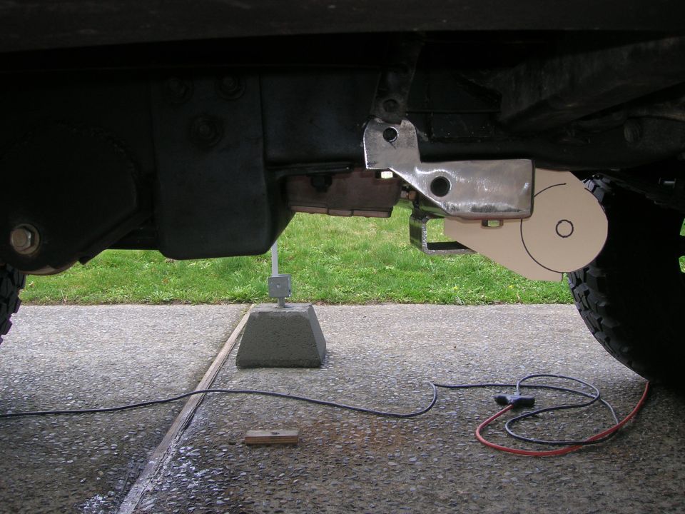 Close-up of a metal bracket attached to a vehicle's frame, with a circular white component nearby. Wires and a concrete block are on the ground