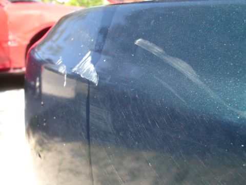 1995 Ford Taurus - Scratched dark green car bumper with another vehicle in the background.