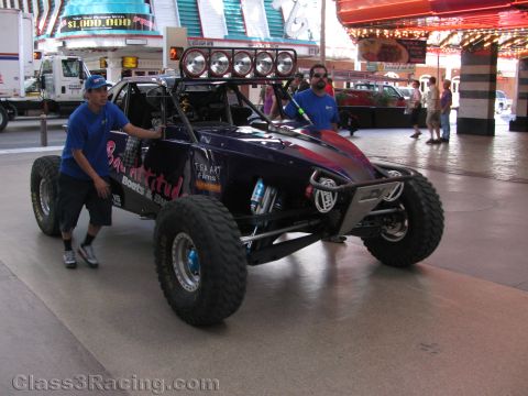 1990 Ford Bronco - man in blue pushes a purple-and-red open desert race buggy with a roof light bar along a covered Las Vegas walkway.