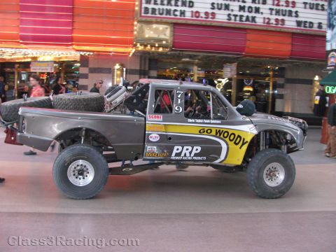 1990 Ford Bronco - gray off-road race truck numbered 719 with GO WOODY and PRP graphics passes beneath a lit casino marquee.