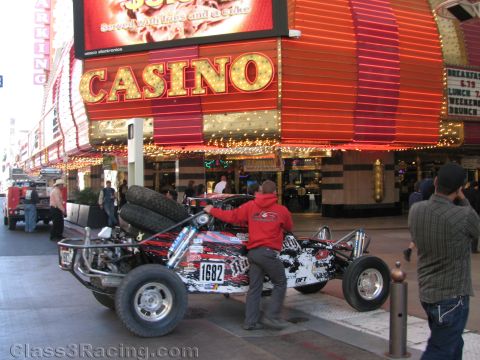 1990 Ford Bronco - man in red rolls a black graphic-wrapped off-road race buggy numbered 1682 beneath a glowing CASINO sign in Las Vegas.