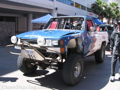 1990 Ford Bronco - blue off-road race truck numbered 311 with a roof light bar and big tires sits in front of a parking structure and palm trees.