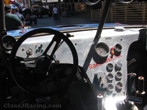 1990 Ford Bronco - View inside an off-road race truck cab showing a bare aluminum dash, round gauges, switches and a black steering wheel.