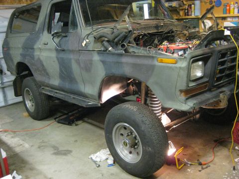 1979 Ford Bronco - Camouflaged Ford Bronco undergoing work in a garage, showing its engine and suspension components.