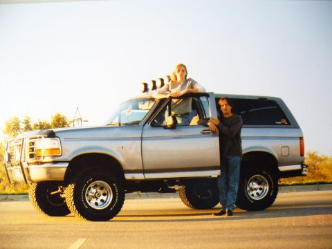 1994 Ford Bronco - A silver Ford Bronco is parked on the side of the road with two people posing next to it.