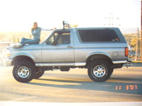 1994 Ford Bronco - A silver SUV with a woman posing on its hood is seen from the side, with a timestamp of 11:30'7 visible.
