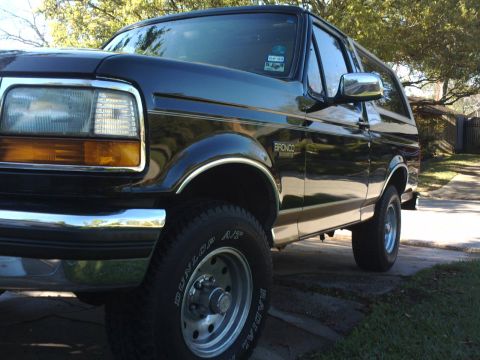 1994 Ford Bronco - A black Ford Bronco is parked on a paved surface, with trees and a fence visible in the background.
