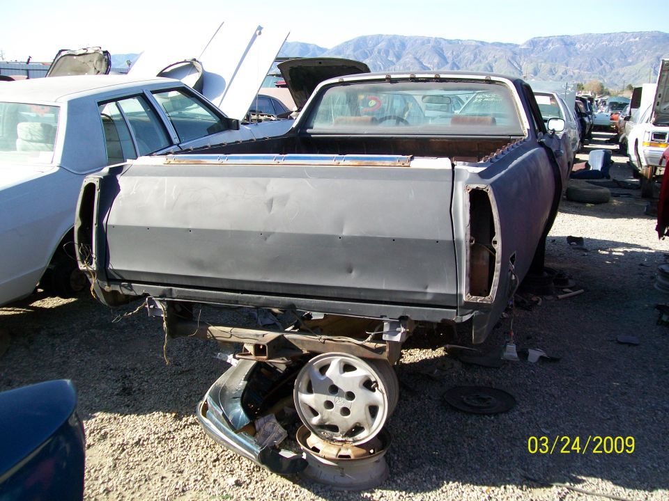 Rear of a stripped-down pickup truck in a junkyard, with another car to its left