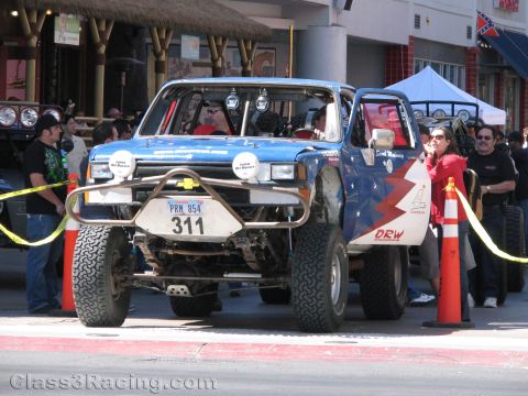 1990 Ford Bronco - blue off-road race truck numbered 311 sits behind yellow caution rope and orange cones with spectators alongside.