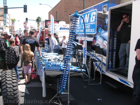 1990 Ford Bronco - King Off-Road Racing Shocks trailer display with rows of blue coilover shock absorbers as a crowd browses the street fair.