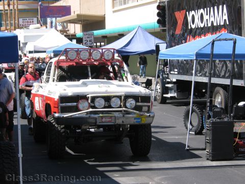 1990 Ford Bronco - red and white off-road race truck with a roof light bar drives down a street past a Yokohama tire trailer and blue tents.