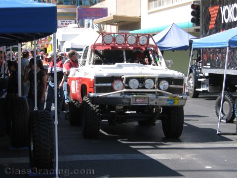 1990 Ford Bronco - white off-road race truck with a roof light bar and big tires sits among vendor tents at a downtown street event.