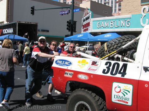 1990 Ford Bronco - Several men push a red and white off-road race truck numbered 304 along a downtown street under a Gambling sign.