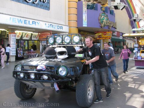 1990 Ford Bronco - smiling man poses beside a black off-road race truck with light bar inside a casino arcade with a neon rainbow above.