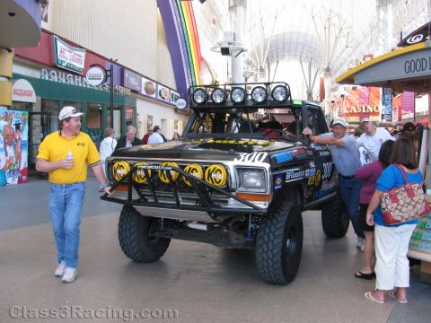 1990 Ford Bronco - black off-road race truck numbered 300 with a roof light bar is pushed through a covered casino pedestrian mall.