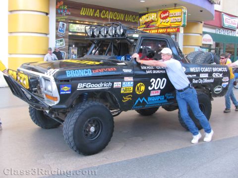 1990 Ford Bronco - black off-road race truck numbered 300 lettered West Coast Broncos with a roof light bar parked at a street event.