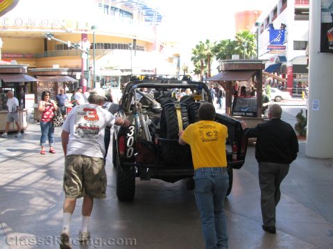 1990 Ford Bronco - Three men push a black off-road race truck numbered 300 from behind through a downtown casino pedestrian mall.