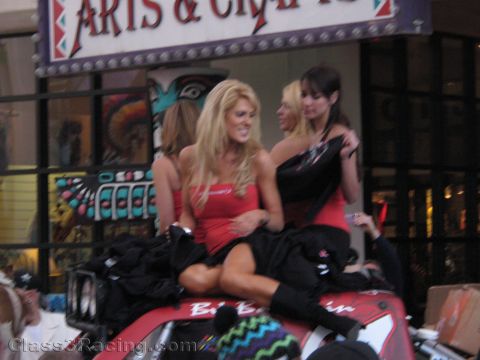 1990 Ford Bronco - Several women in red promotional outfits pose seated on a red race vehicle beneath an Arts and Crafts storefront sign.