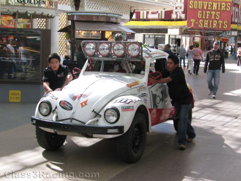 1990 Ford Bronco - Two men push a white Volkswagen Beetle baja bug fitted with a roof light bar through a covered casino pedestrian mall.