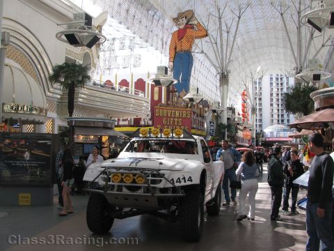 1990 Ford Bronco - white off-road race truck numbered 1414 sits in a covered casino mall beneath the giant neon cowboy sign as people walk by.