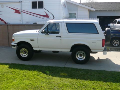 1988 Ford Bronco - Driver-side profile of a white two-door Ford Bronco full-size SUV with matching white hardtop, parked on a concrete driveway beside a lawn.…