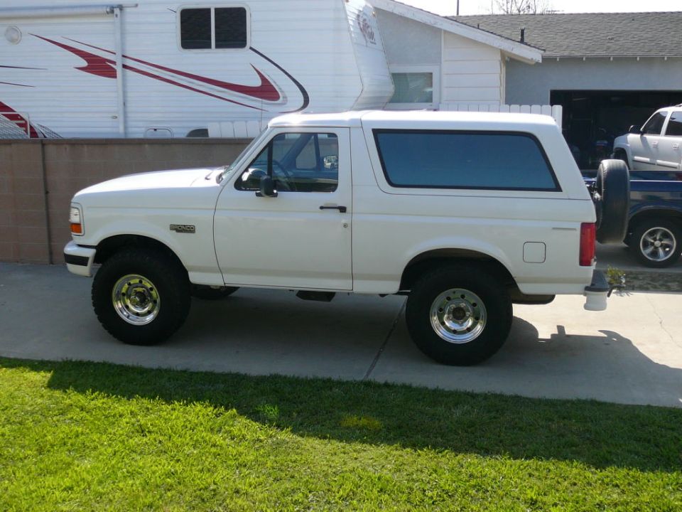 White two-door Ford Bronco, driver-side profile, on chrome wheels and all-terrain tires in a driveway