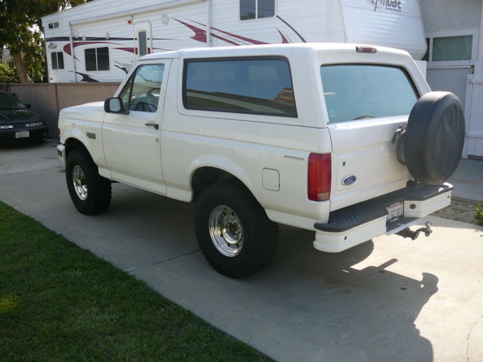 White Ford Bronco parked on driveway, rear-mounted spare tire, trailer in background