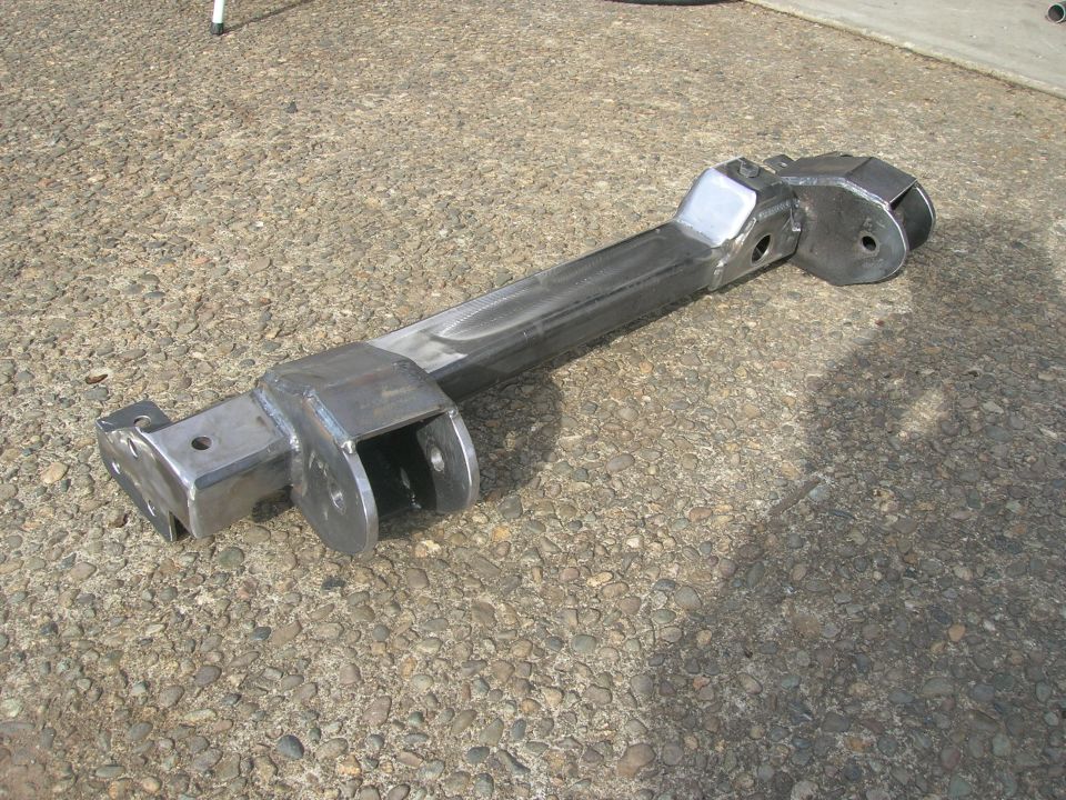 Fabricated metal suspension arm with multiple mounting points and pivot holes, resting on a gravel surface