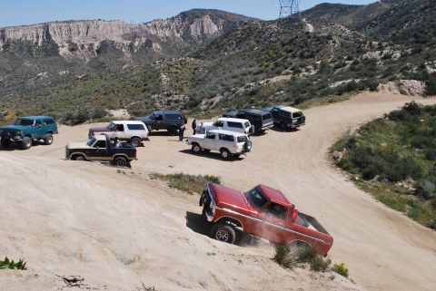 1996 Ford Bronco - Several Ford Broncos are gathered on a dusty, off-road trail in a mountainous area.
