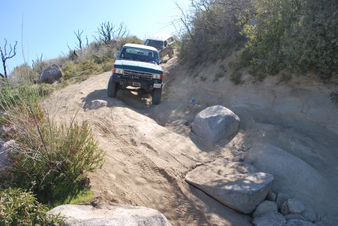 1996 Ford Bronco - Two SUVs navigate a challenging off-road trail with large rocks and sparse vegetation.