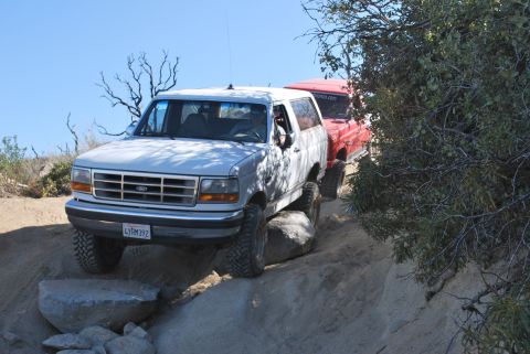 1996 Ford Bronco - A white 1996 Ford Bronco tackles an off-road obstacle course, followed closely by another vehicle.