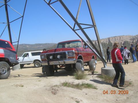 1985 Ford Bronco - A lifted red Ford truck is parked near a power line tower with other vehicles and people present.