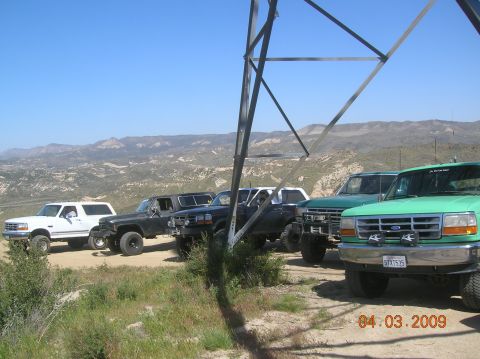 1985 Ford Bronco - Several Ford trucks and a Bronco are parked on a dirt road with a mountainous landscape in the background.