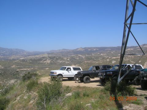 1985 Ford Bronco - A white Ford Bronco is parked on a dirt road with other vehicles, overlooking a vast, hilly landscape under a clear blue sky.