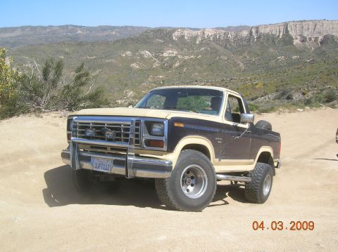 1985 Ford Bronco - This two-tone Ford Bronco is parked on a dusty trail with a scenic mountain range behind it.