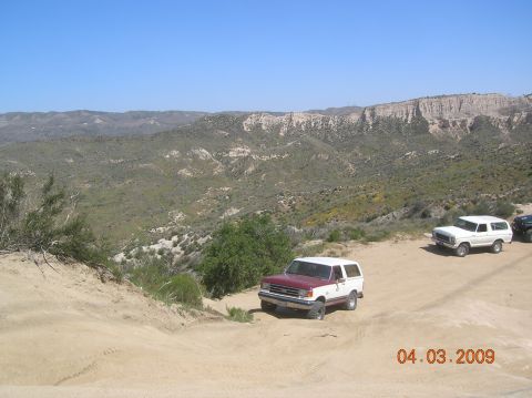1985 Ford Bronco - Two Ford Broncos and a black vehicle are parked on a dirt road with a mountainous landscape in the background.