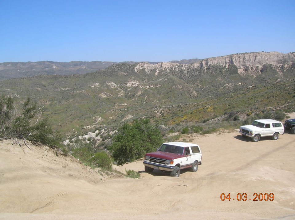 Maroon and white Ford Bronco on a dirt road, with a white Ford Bronco and a black vehicle behind it