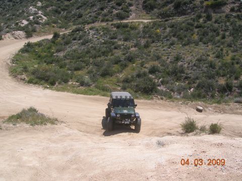 1985 Ford Bronco - A green Jeep Wrangler navigates a dirt road in a dry, hilly terrain.