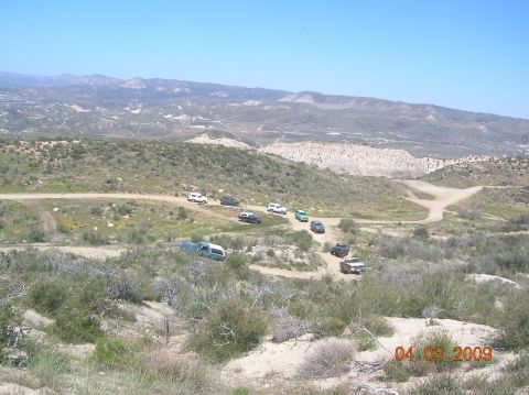 1985 Ford Bronco - Several vehicles traverse a dusty, winding path through a dry, mountainous terrain under a clear blue sky.