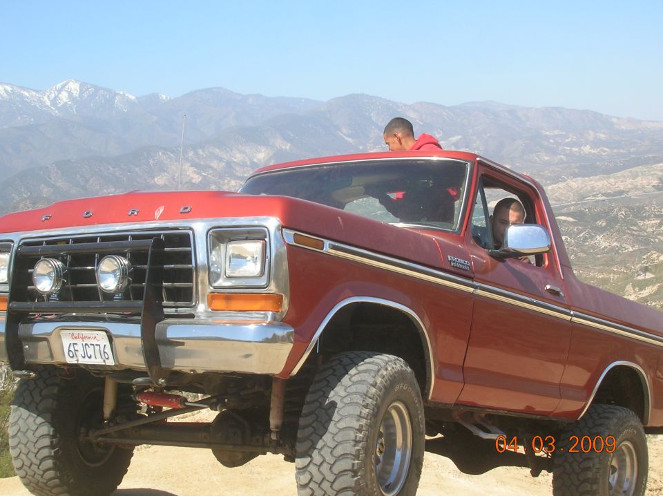 Red Ford Bronco with off-road tires and a brush guard, parked on a dirt road with mountains in the background