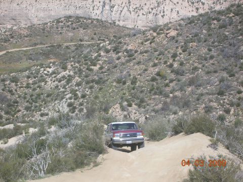 1985 Ford Bronco - A red and white SUV navigates a dusty trail through a dry, hilly landscape.