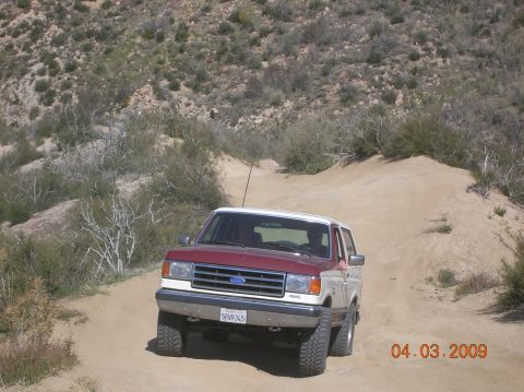 1985 Ford Bronco - Ford Bronco navigates a dusty, off-road trail with sparse vegetation and hills in the background.