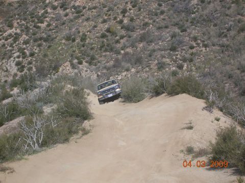 1985 Ford Bronco - A truck navigates a dusty, off-road trail in a dry, scrubby landscape.