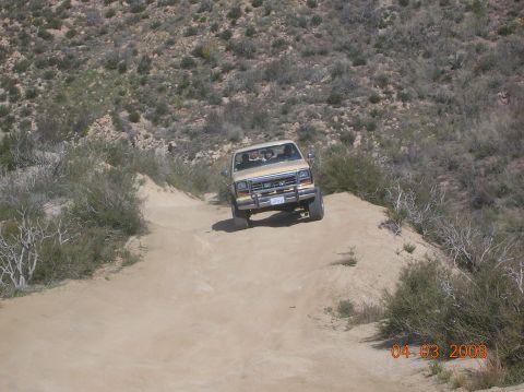 1985 Ford Bronco - A tan SUV navigates a dusty trail in a dry, hilly landscape.