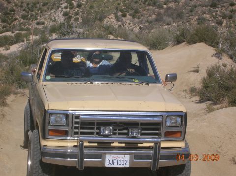 1985 Ford Bronco - This tan Ford Bronco, likely from the early 1980s, is seen off-road with a prominent grille guard and auxiliary lights.