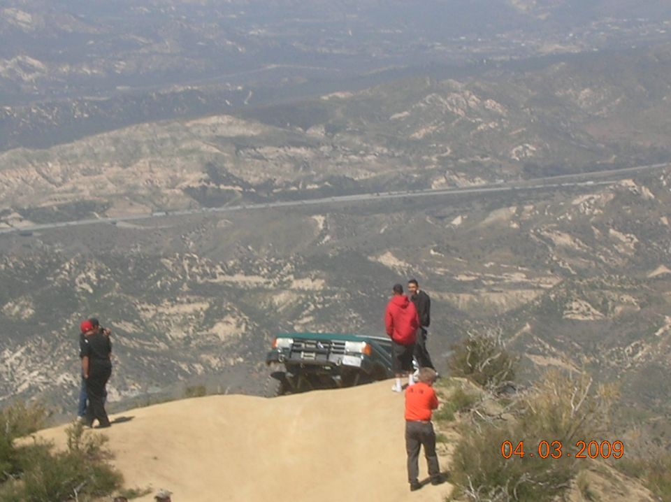 Green off-road vehicle with people standing around it on a dirt cliff edge, overlooking a valley
