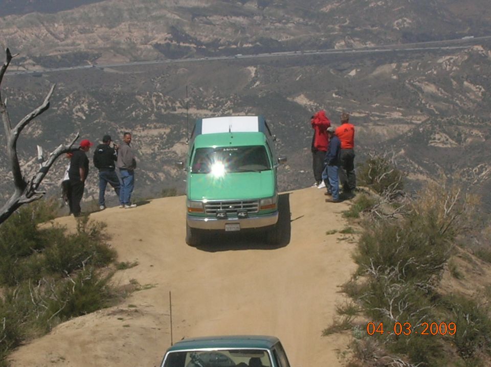 Green Ford truck on dirt road with people standing on the edge of a cliff. Another vehicle is visible below