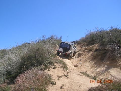 1985 Ford Bronco - Multiple off-road vehicles navigate a steep, dusty trail under a clear blue sky.