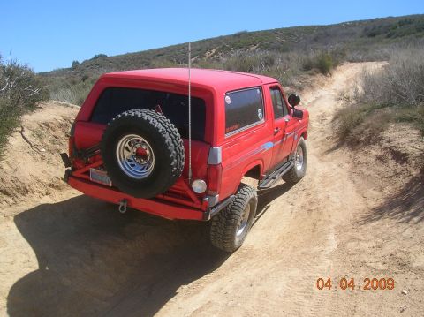 1985 Ford Bronco - Red Ford Bronco navigates a narrow dirt path with scrub brush and hills in the background.