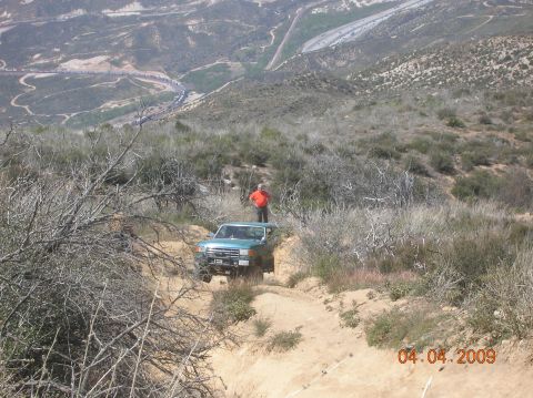 1985 Ford Bronco - A person stands atop a teal SUV as it navigates a , off-road trail with a scenic mountainous landscape in the background.